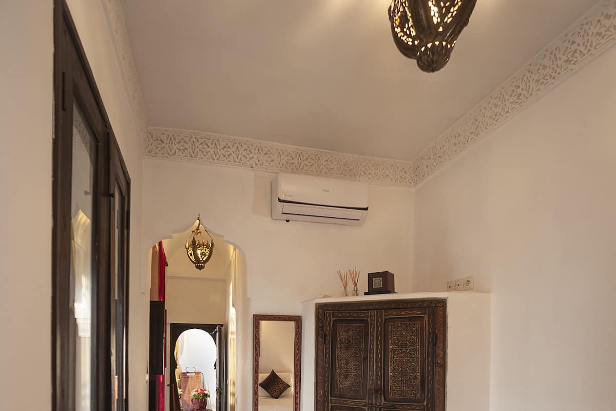 A hallway with a patterned rug, a bed with a white bedspread, a wooden wardrobe, and a hanging light fixture.