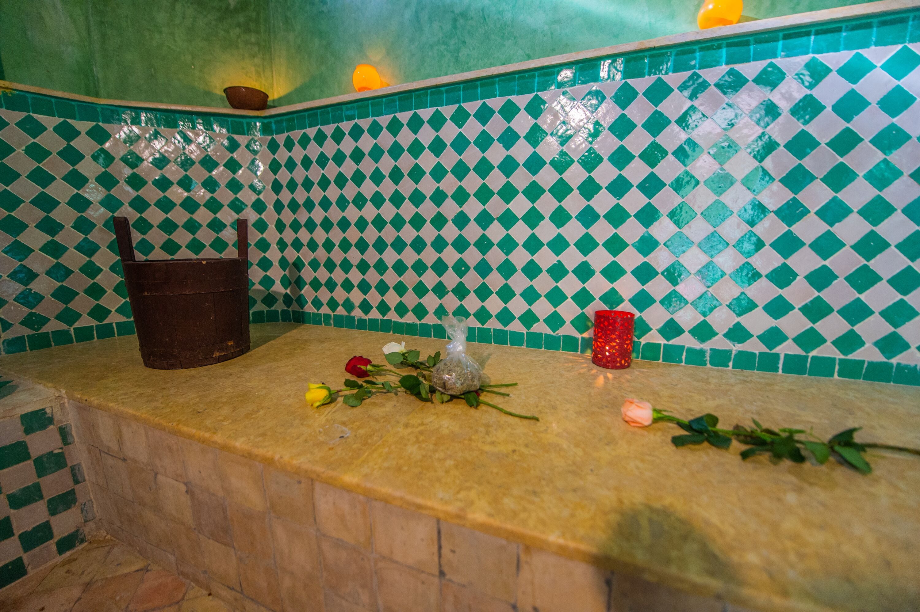 Un baño con salpicadero de azulejos a cuadros, un lavamanos y una encimera con una botella, un vaso rojo y flores.