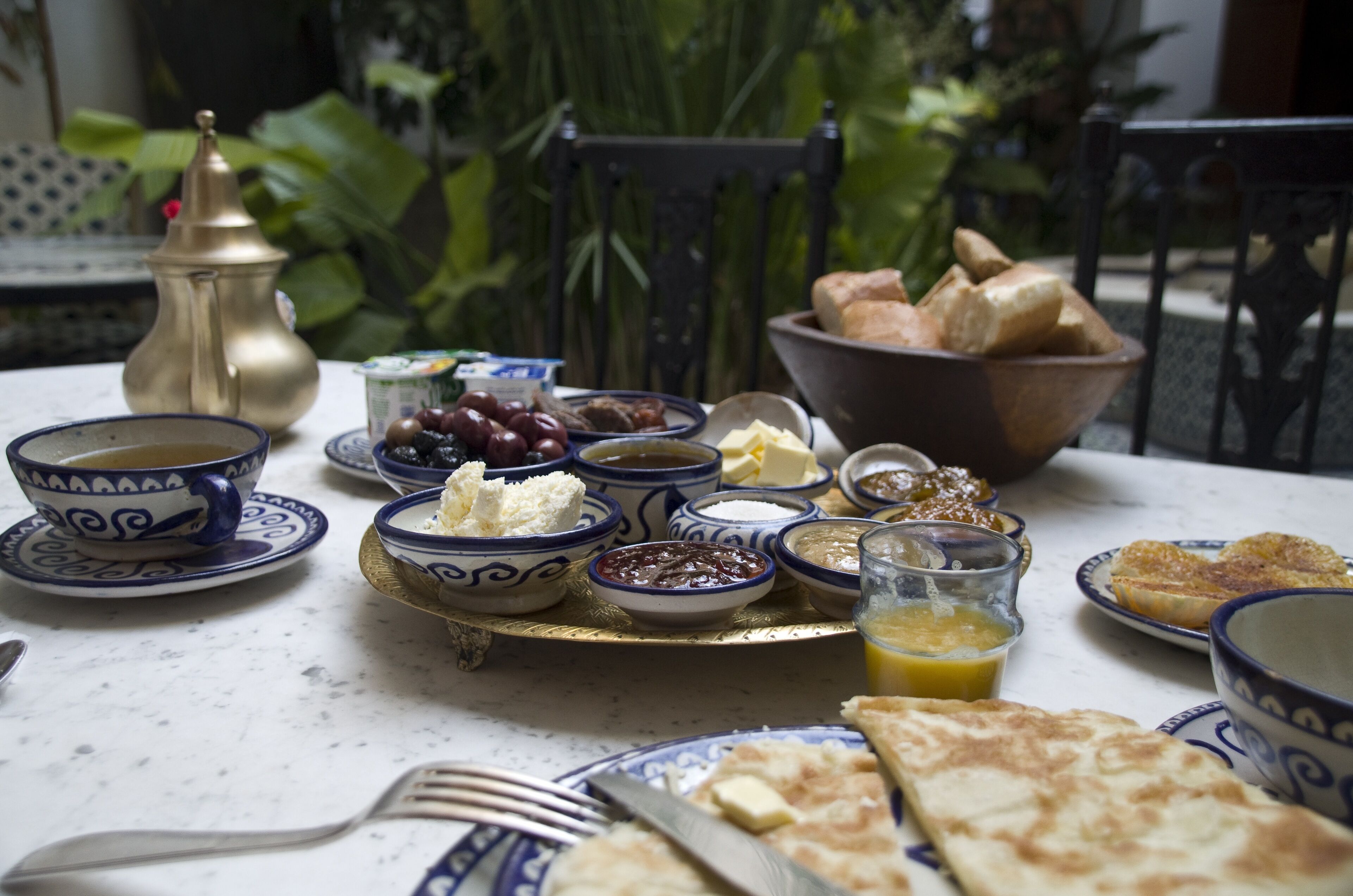 Una mesa servida con una variedad de platos, incluyendo pan, aceitunas y queso, junto con una tetera de bronce y cuencos sobre un mantel blanco.