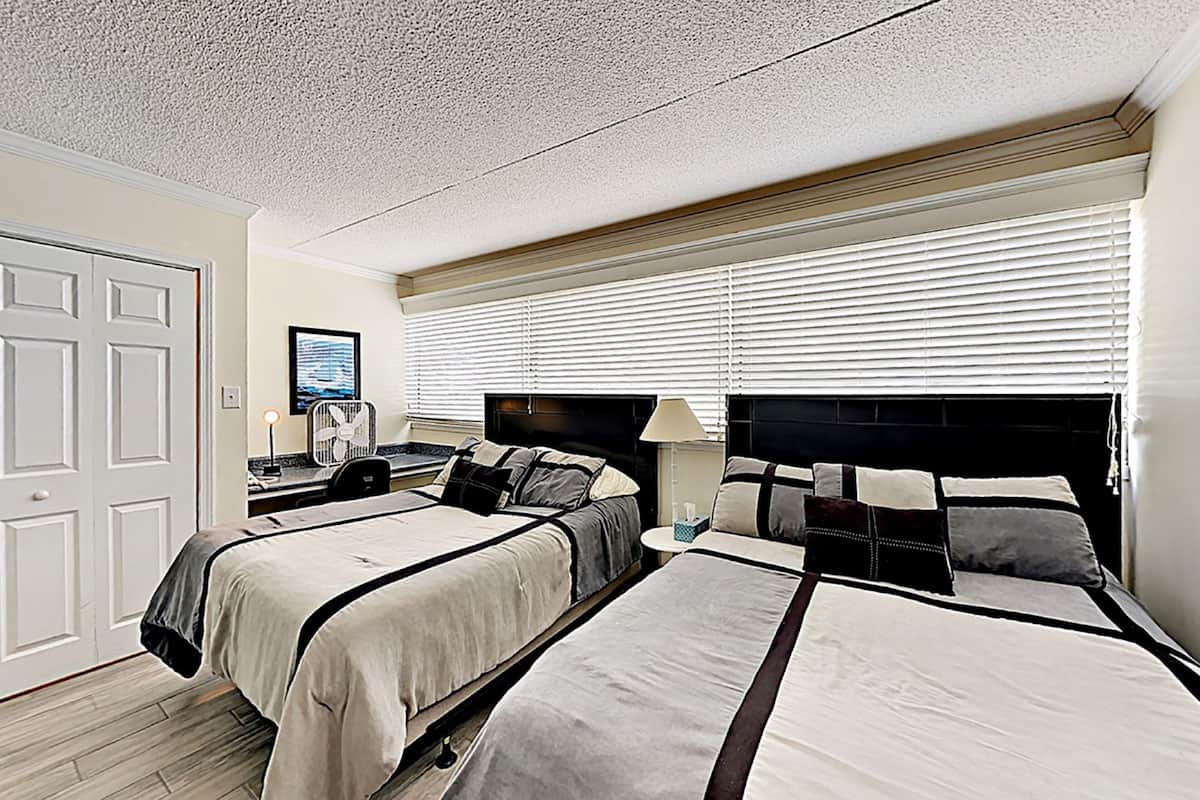 A hotel room with two beds, a desk, a chair, a lamp, and a window with blinds.