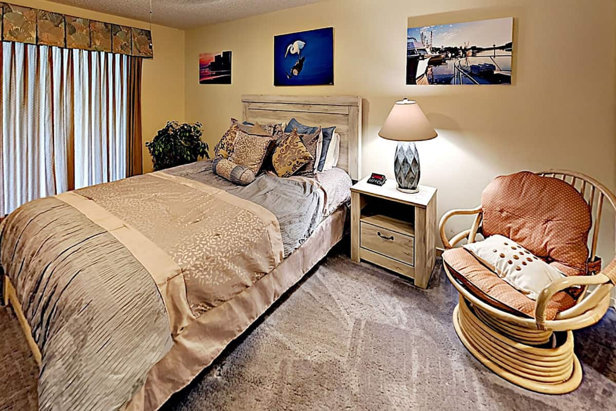 A bedroom with a large bed, a nightstand with a lamp, a wicker chair, and two wall-mounted paintings.