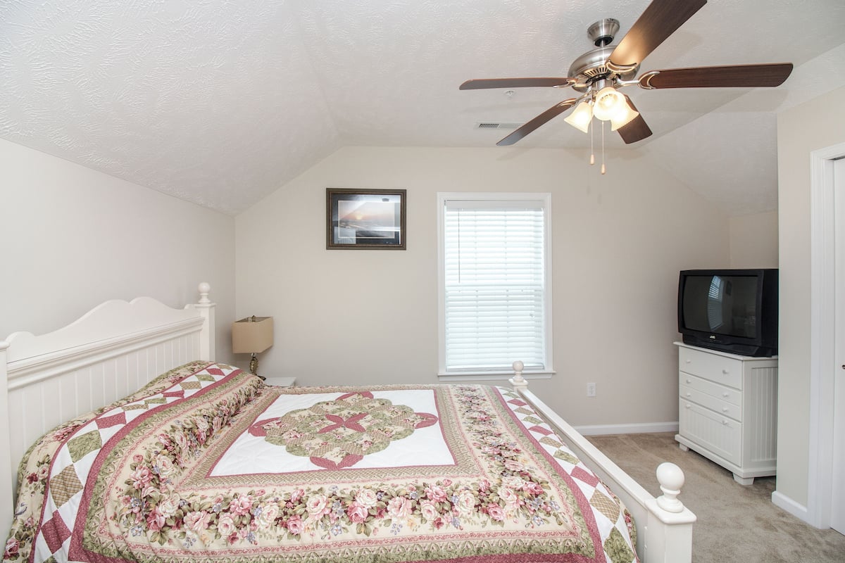A bedroom with a bed, a ceiling fan, a window with blinds, a dresser, and a television.