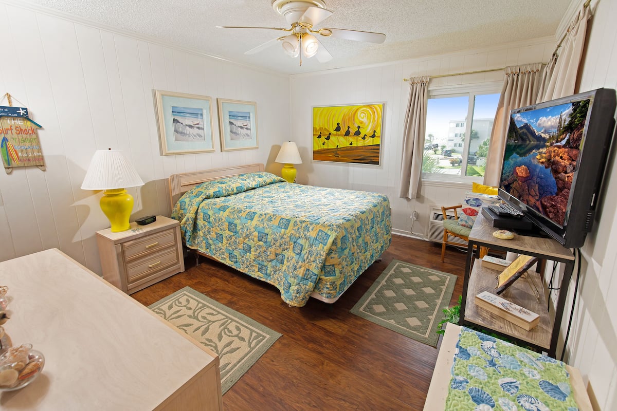 A bedroom with a bed, two bedside lamps, a ceiling fan, a TV, and a window with curtains.