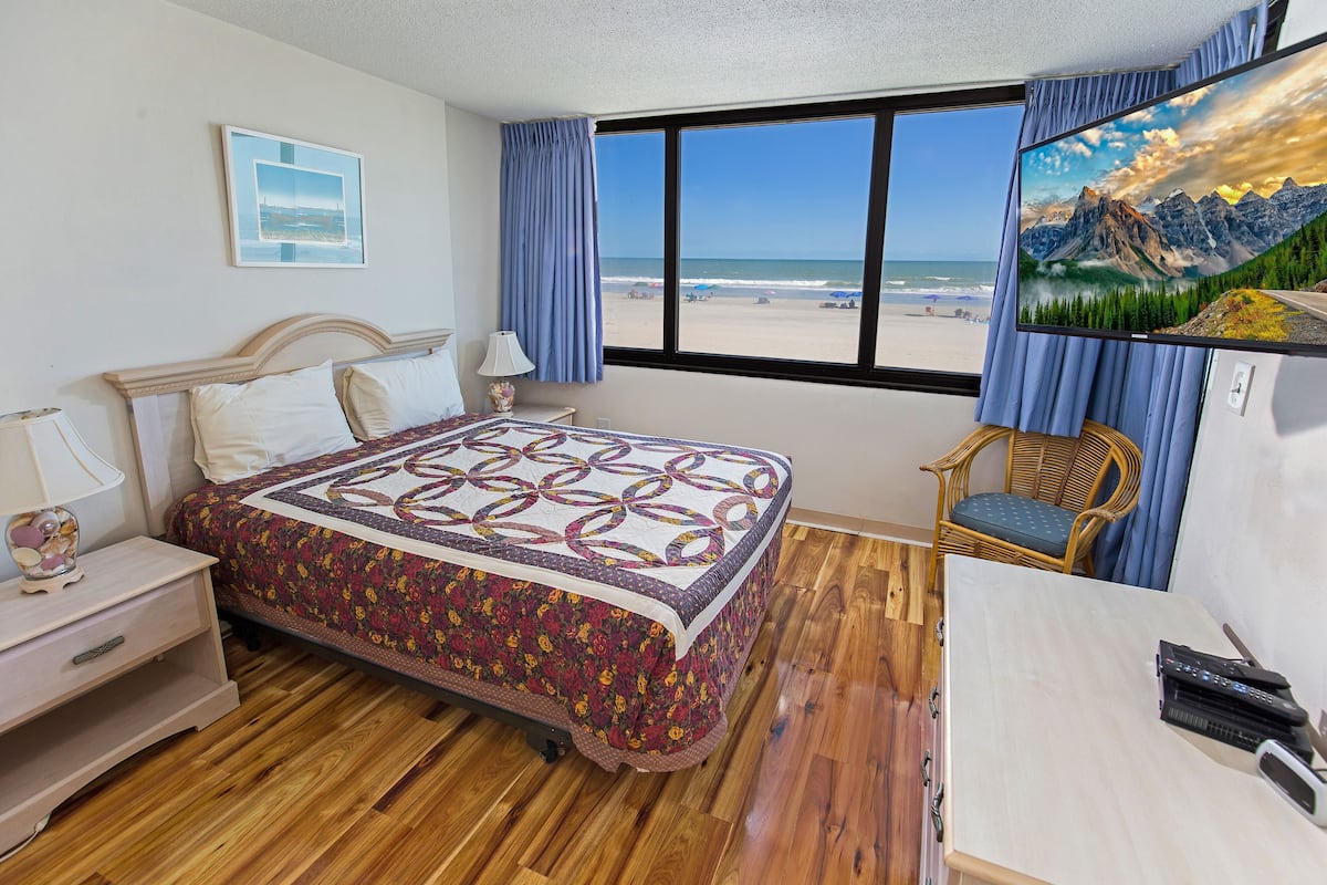A bedroom with a bed, a chair, a desk, and a large window with a beach view.