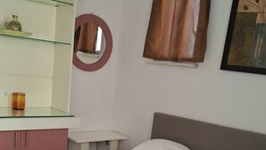 A neatly made bed with towels, a bedside table, a mirror, a window with curtains, and a wall-mounted shelf with items. - Mastic Point Studios (Chios)
