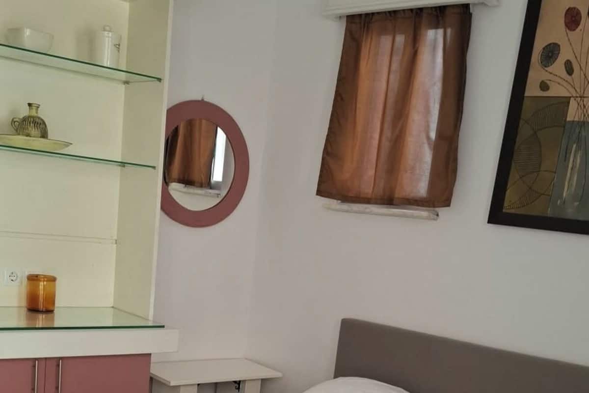 A neatly made bed with towels, a bedside table, a mirror, a window with curtains, and a wall-mounted shelf with items.