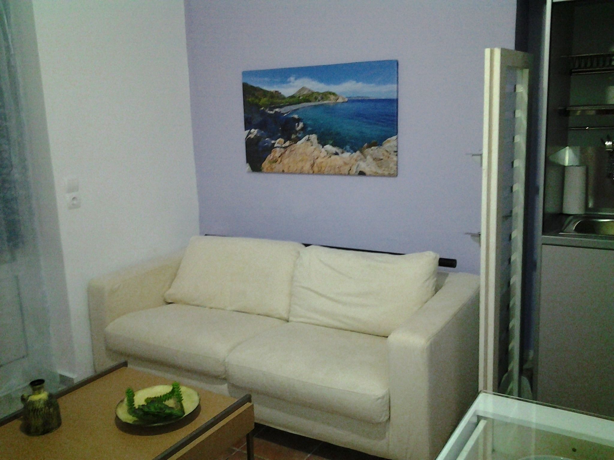 A living room with a beige sofa, a painting of a coastal landscape, and a kitchen area visible through an open door.
