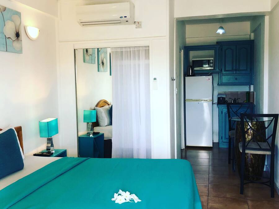 Habitación de hotel con cama, mesita de noche, lámpara, espejo, kitchenette y zona de comedor.