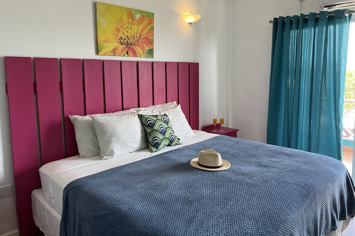 Un dormitorio con cabecera rosa, una cama con ropa de cama blanca, una manta azul, un cojín verde estampado y un sombrero sobre la cama. Hay un cuadro de una flor en la pared y una mesita de noche con dos vasos de bebida.