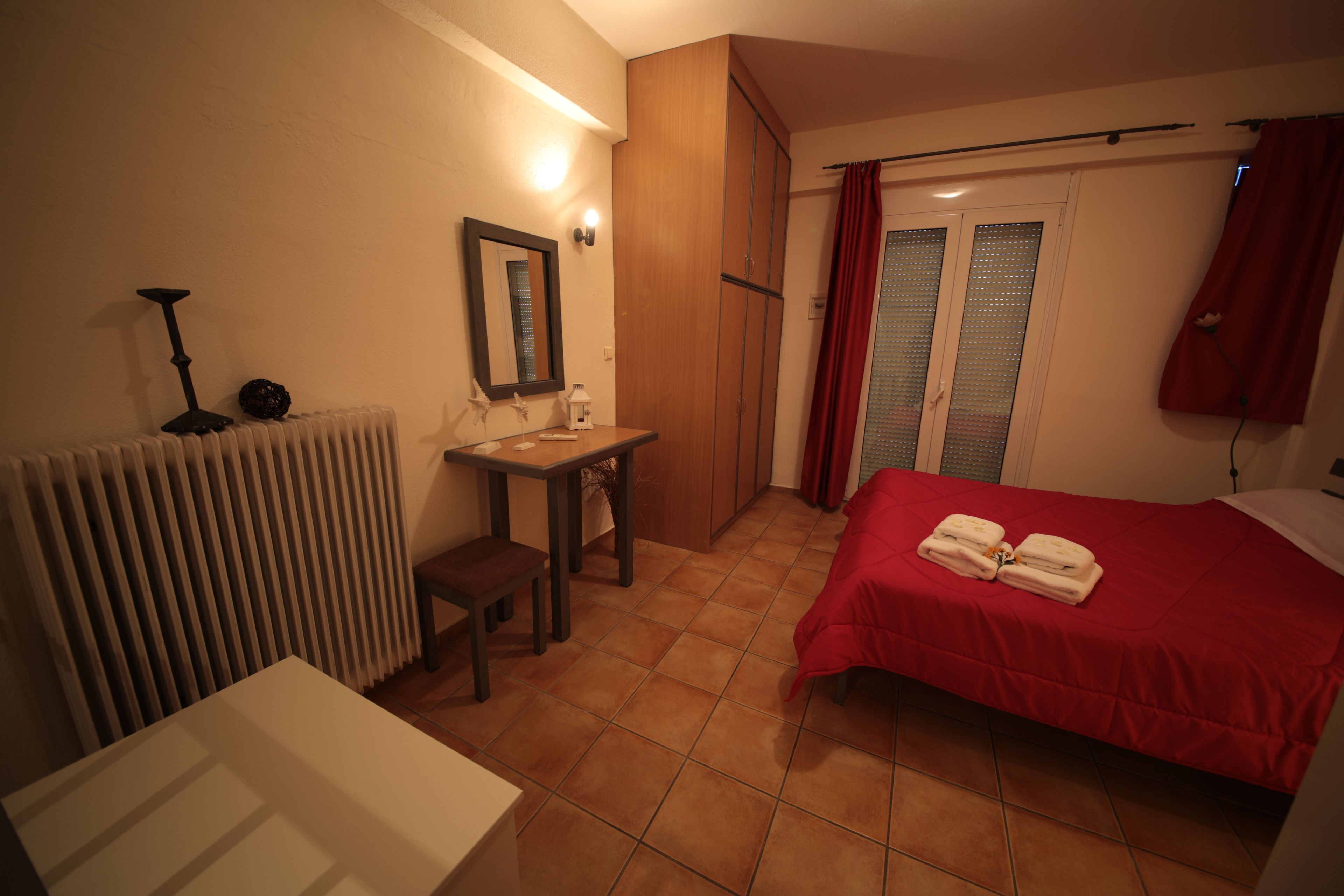 A hotel room with a bed, a desk, a chair, a radiator, a mirror, and a window with curtains.