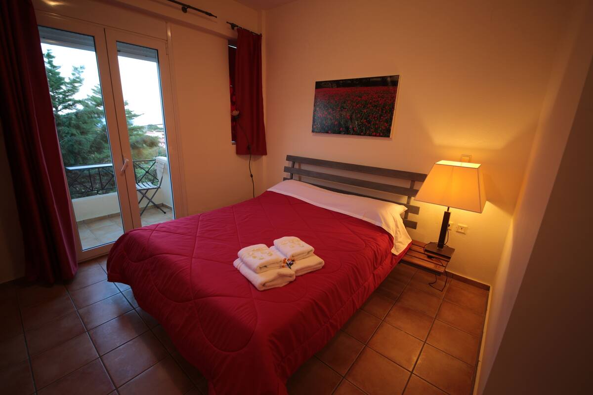 Ein Schlafzimmer mit roter Bettdecke, einem Nachttisch mit Lampe, einem Bild an der Wand und Blick durch eine Schiebetür auf einen Außenbereich.