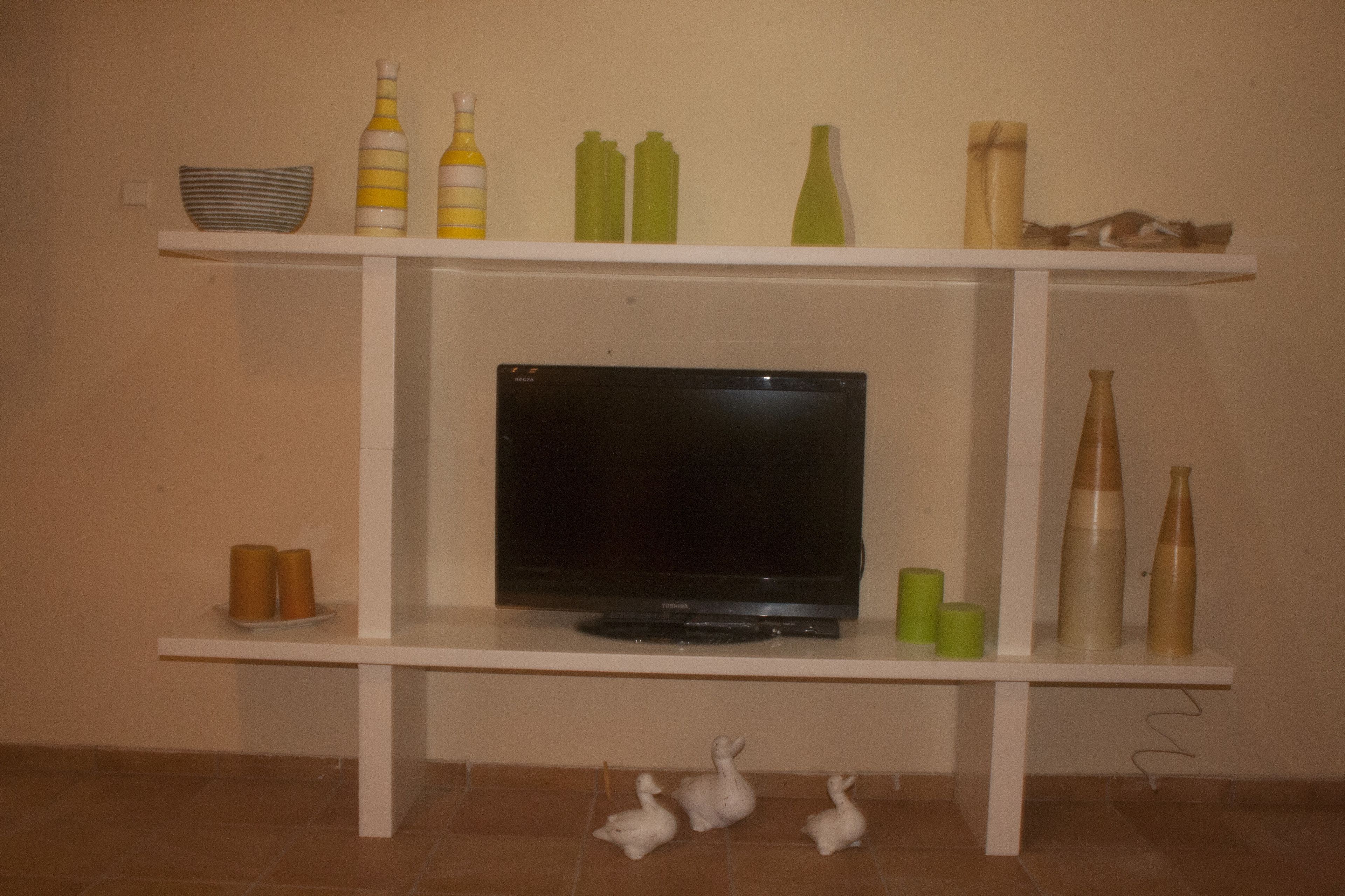 Un televisor sobre un pedestal blanco con repisas arriba y abajo, que exhiben varios objetos decorativos.