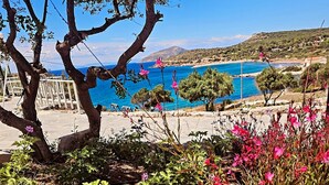 A garden with various plants and flowers, a tree, and a view of a blue sea and distant hills. - Poseidon Beach Villas (Lavreotiki)
