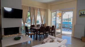 A spacious living area with a dining table, a sofa, a flat-screen TV, and a view of the sea through large windows. - Poseidon Beach Villas (Lavreotiki)