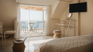 A bedroom with a bed, a TV, a window with curtains, a chair, and a table with a view of the sea. - Poseidon Beach Villas (Lavreotiki)