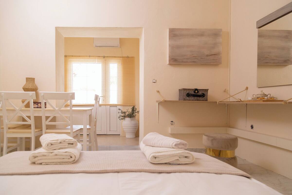 A hotel room with a bed, a desk, and a chair. There is a window with blinds, a potted plant, and a shelf with a basket and a small table.