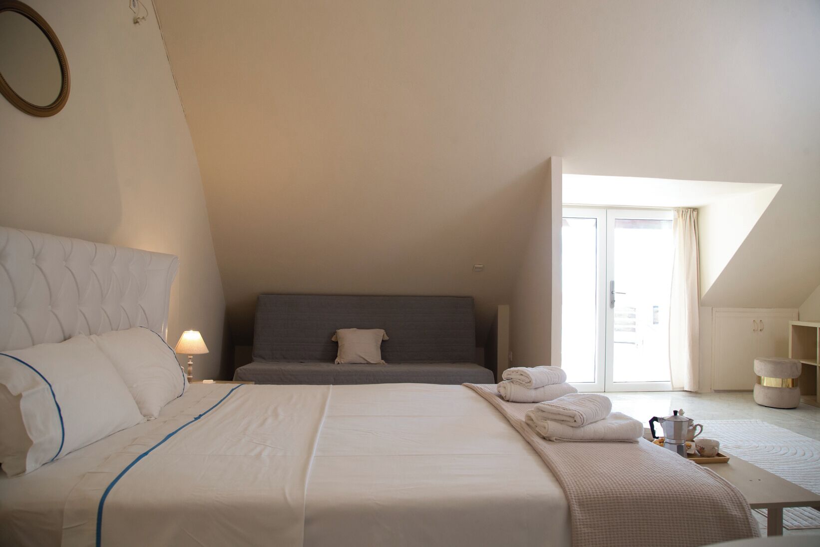 Una camera da letto con un letto grande, un divano più piccolo, un tavolo con asciugamani e un bollitore, e vista all'esterno attraverso una finestra.