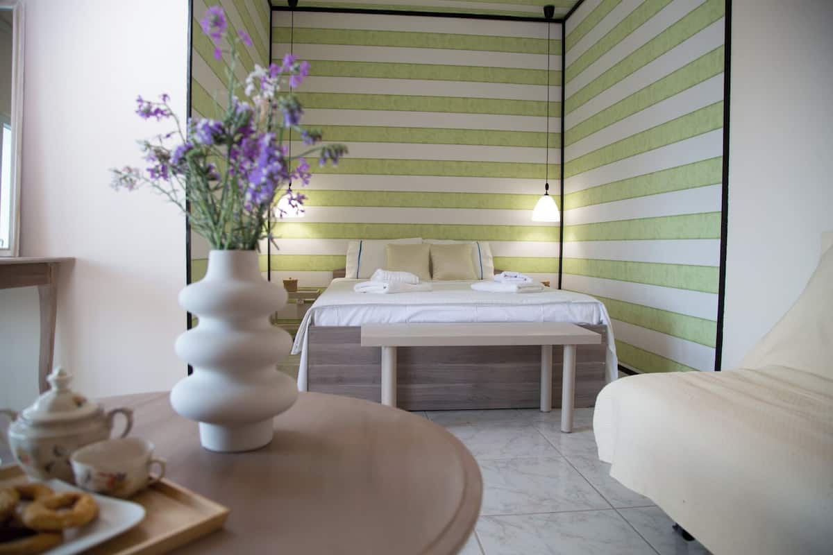 A hotel room with a bed, a bedside table with a vase of flowers, a chair, and a tray with a teapot and cups.