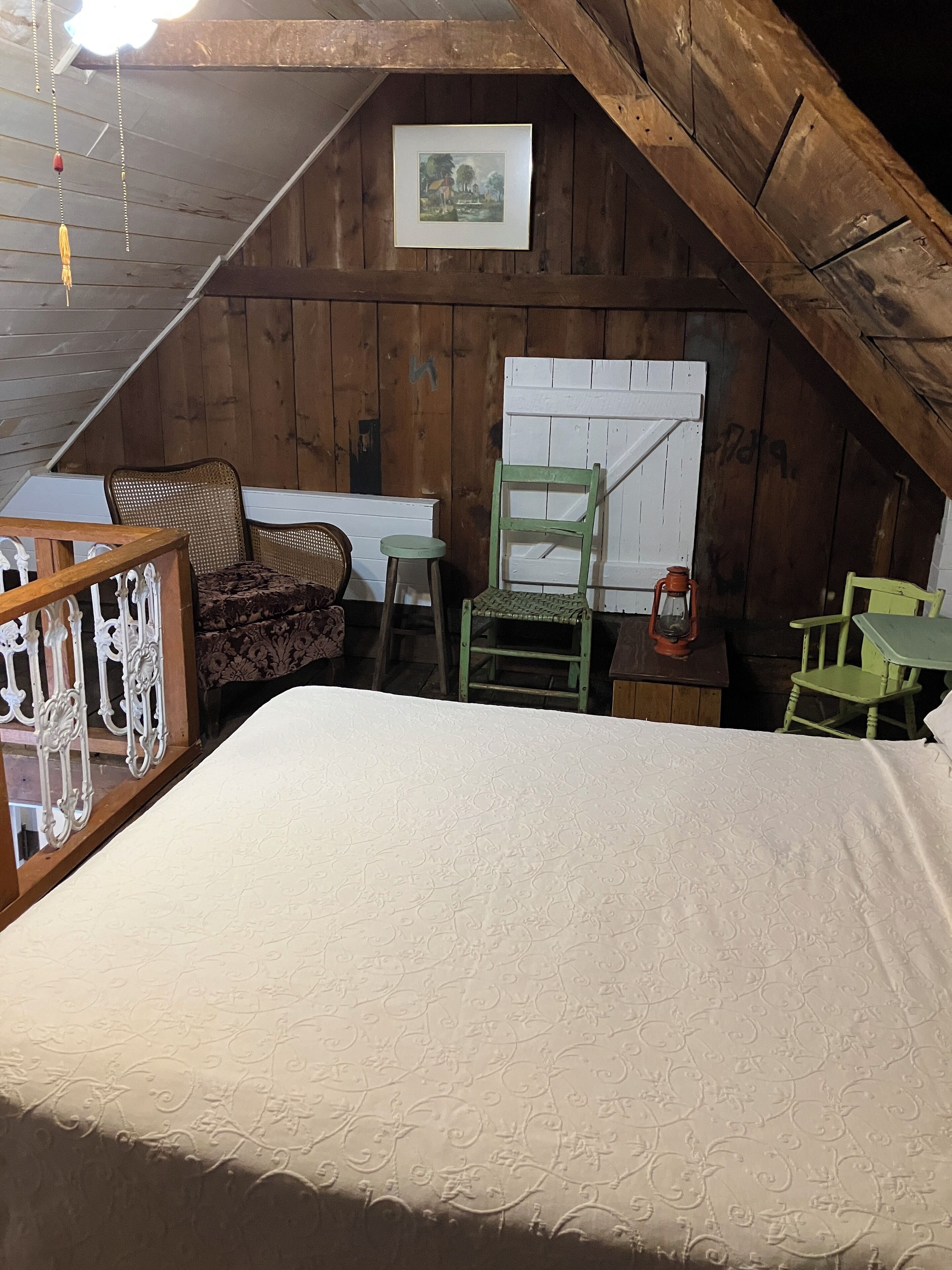 A rustic attic bedroom with a wooden sloped ceiling, a bed, a wicker chair, a green chair, and a small table with a lantern.