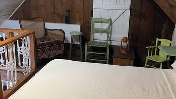 A rustic attic bedroom with a wooden sloped ceiling, a bed, a wicker chair, a green chair, and a small table with a lantern.