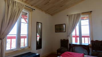 Ein Zimmer mit Holzboden, zwei mit Vorhängen drapierten Fenstern, einem Bett mit roter Decke, einem hölzernen Schminktisch, einem Stuhl und einem gerahmten Bild an der Wand.