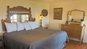 A bedroom with a large bed, wooden headboard, two bedside tables, a dresser, a mirror, and a wall-mounted lamp. - Auberge Pourvoirie Isle-aux-Grues (Saint-Antoine-de-l'Isle-aux-Grues)