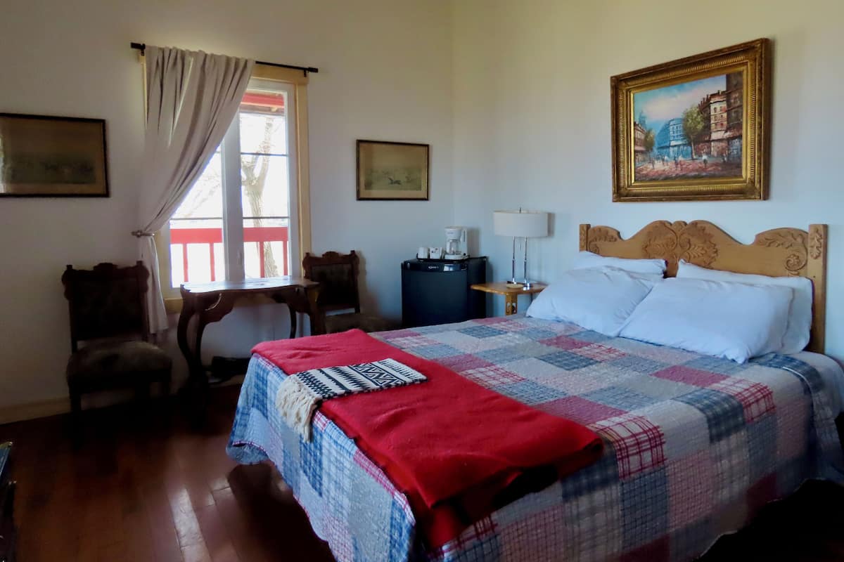 Une chambre d’hôtel avec un lit, un bureau, une chaise, une fenêtre avec des rideaux et deux tableaux encadrés au mur.