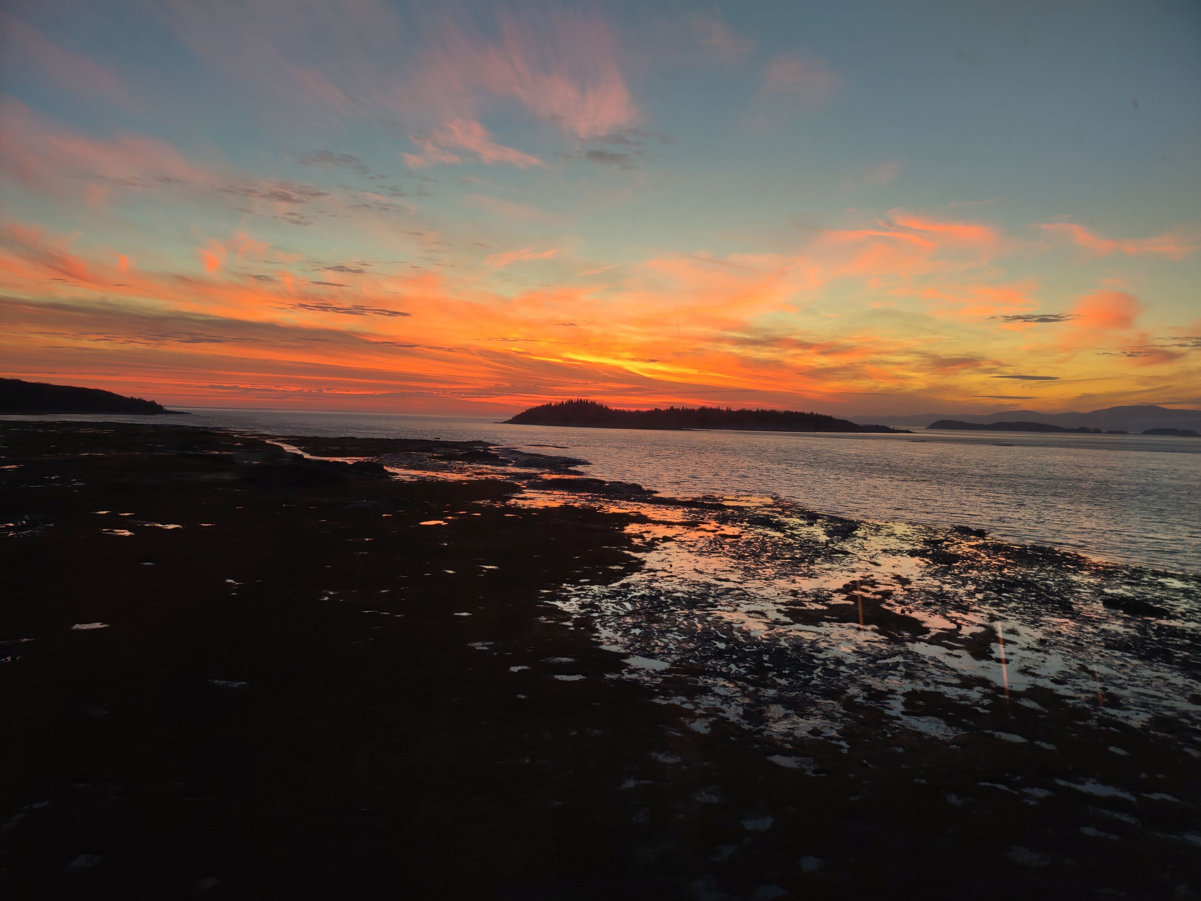 Ein Küsten-Sonnenuntergang mit felsiger Küstenlinie und weiter Landschaft im Hintergrund.