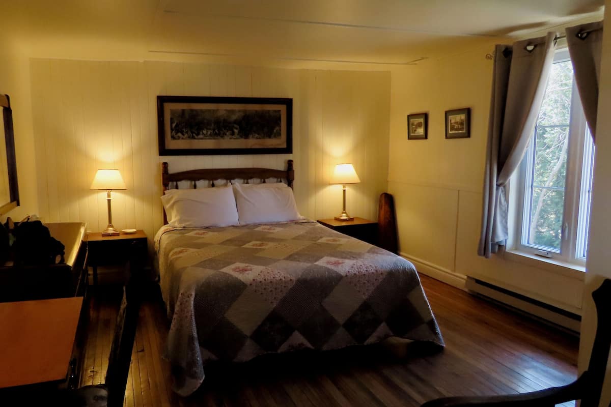 Une chambre à coucher bien rangée, avec un lit, deux lampes de chevet, un tableau encadré au mur et une fenêtre avec des rideaux.