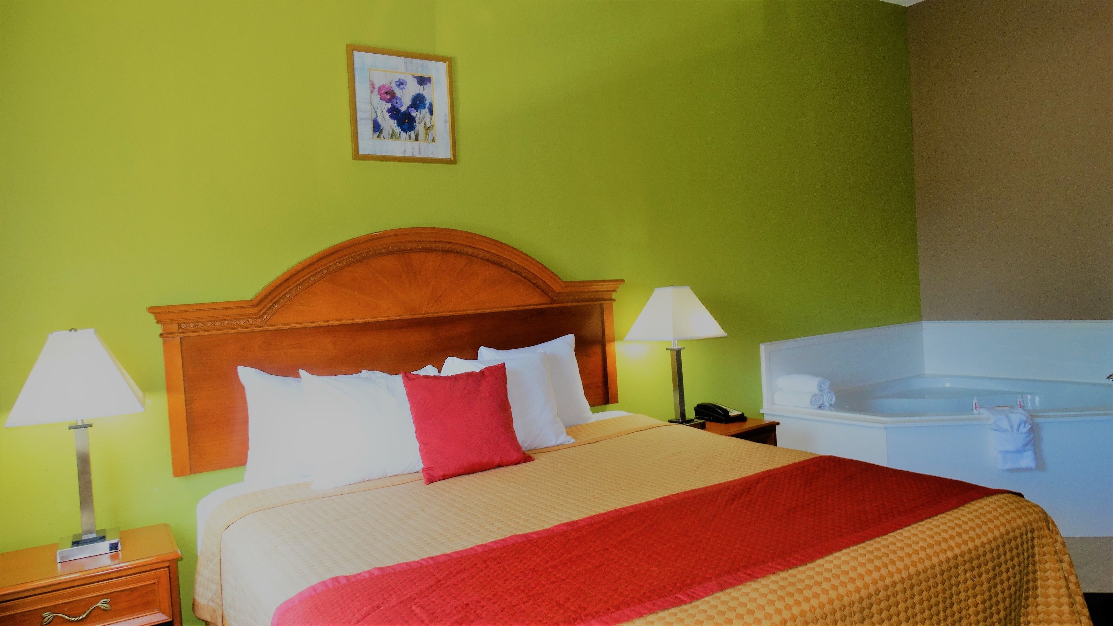 A hotel room with a wooden headboard, a bed with white and red pillows, a red throw blanket, a bedside lamp, a phone, and a bathtub.