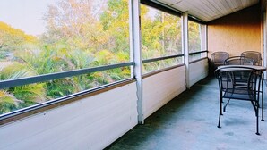 Un balcón con vistas a árboles y plantas, una mesa de metal con sillas y un suelo de hormigón.