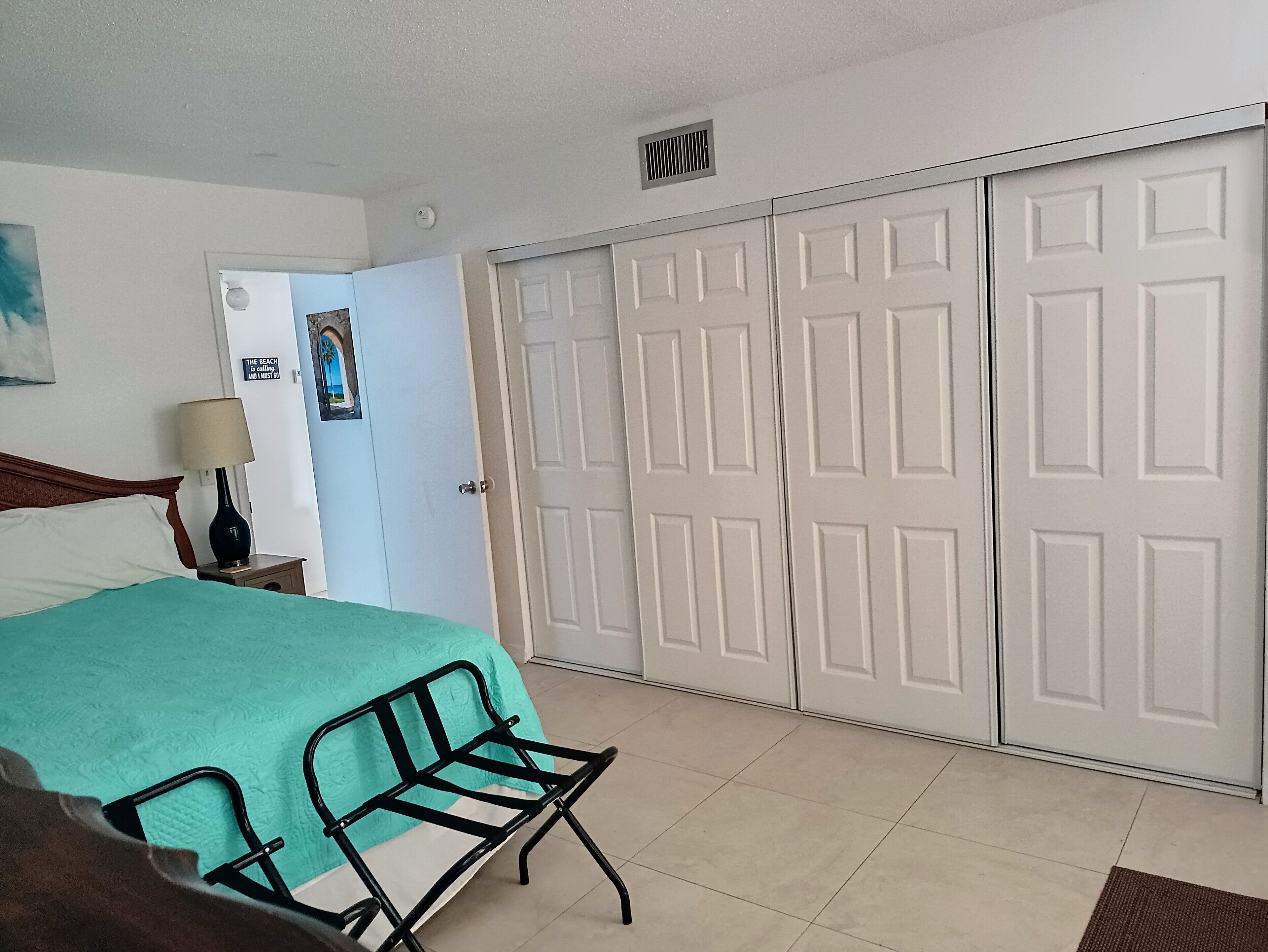Una habitación de hotel con una cama, una silla, un escritorio con lámpara, un armario y una puerta con un cartel.
