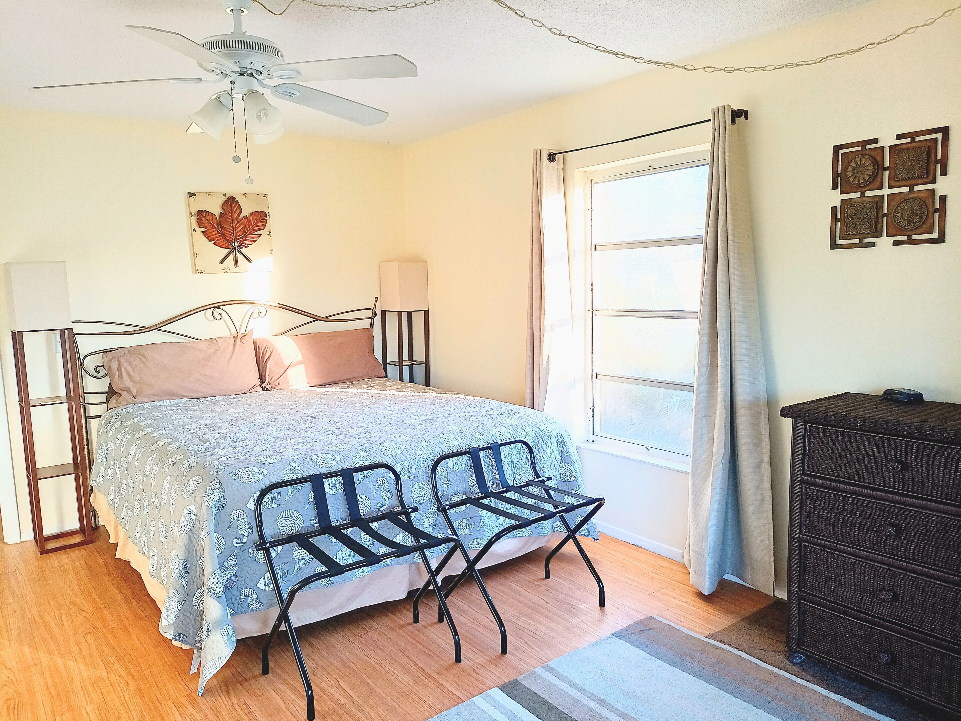 A bedroom with a bed, a ceiling fan, a window with curtains, a dresser, and wall art.