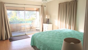 Un dormitorio con una cama grande, un ventilador de techo, una mesita de noche con lámpara y vistas a un balcón con mobiliario exterior.