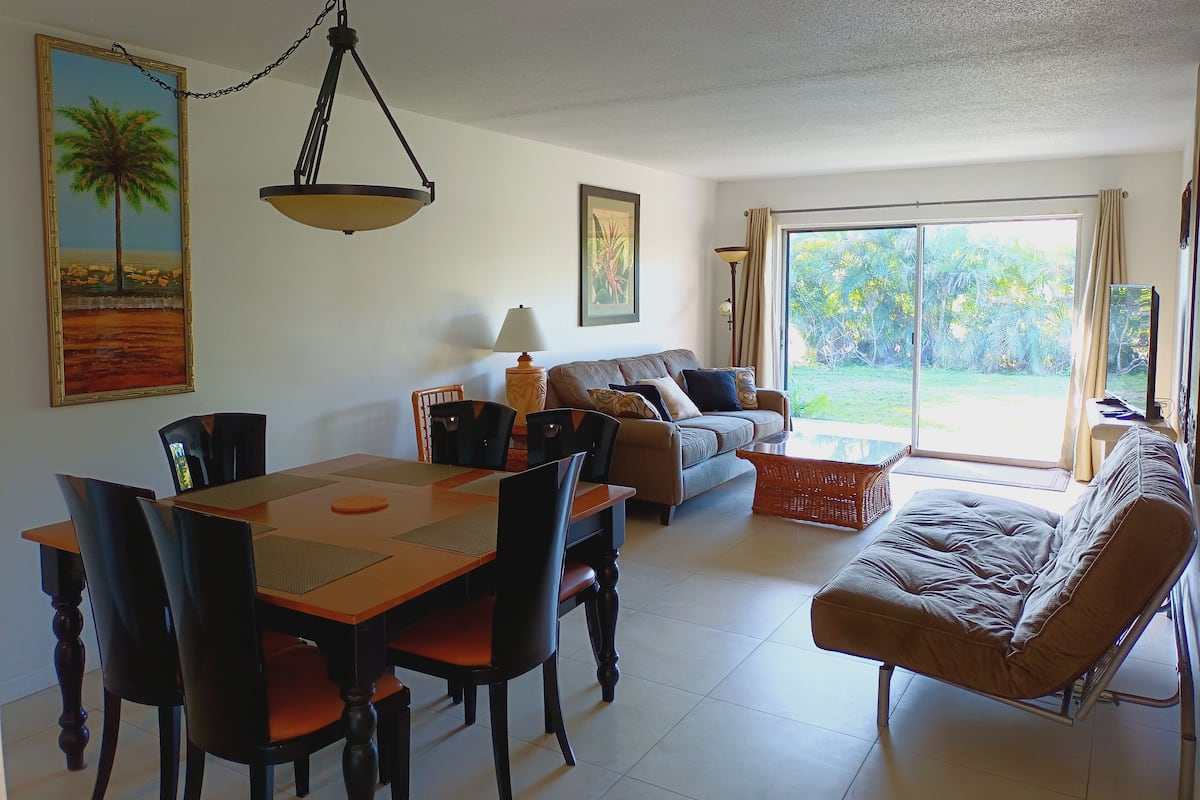 A dining area with a table and chairs, a sofa, and a large window with a view of greenery.