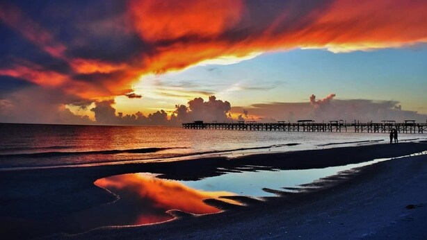 A beach with a pier extending into the water, a sunset in the background, and a person walking on the sand. - Carousel Suites (Redington Shores)
