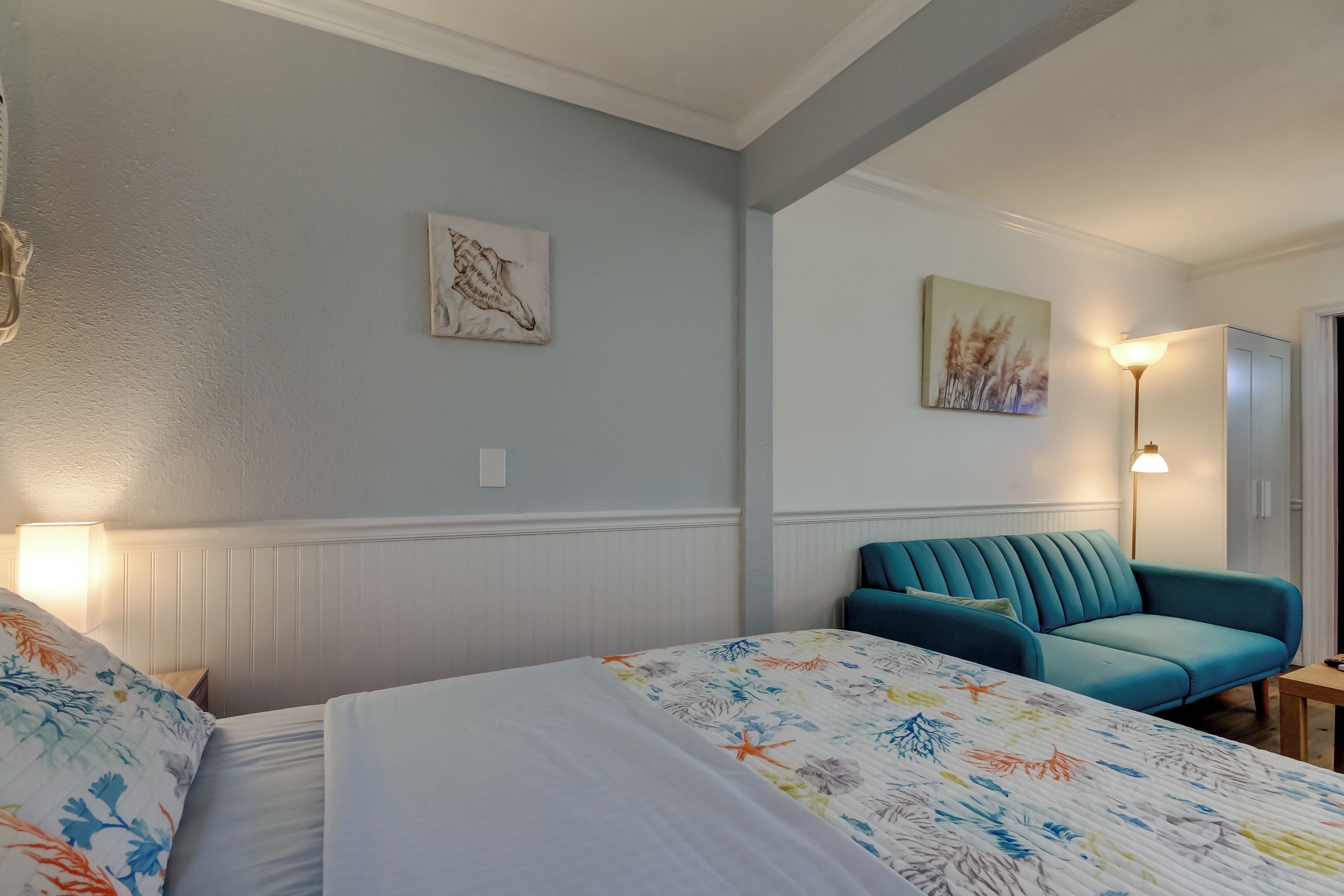 Une chambre d’hôtel avec deux lits, un canapé de couleur turquoise, une lampe et des œuvres d’art accrochées aux murs.