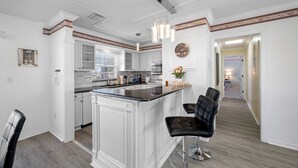 A modern kitchen with white cabinetry, a central island with bar stools, and a decorative clock on the wall. - Carousel Suites (Redington Shores)