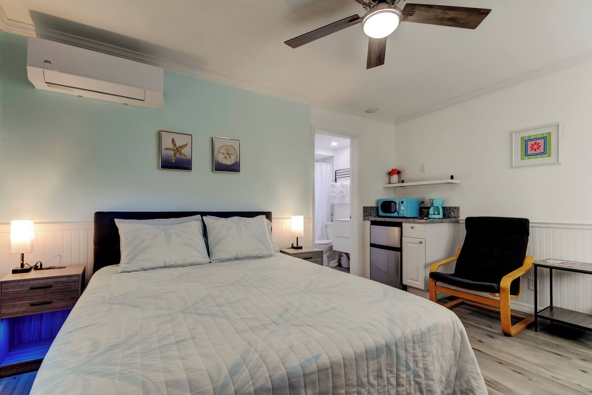 A bedroom with a bed, bedside tables, a ceiling fan, an air conditioning unit, a desk with a chair, and a kitchenette.