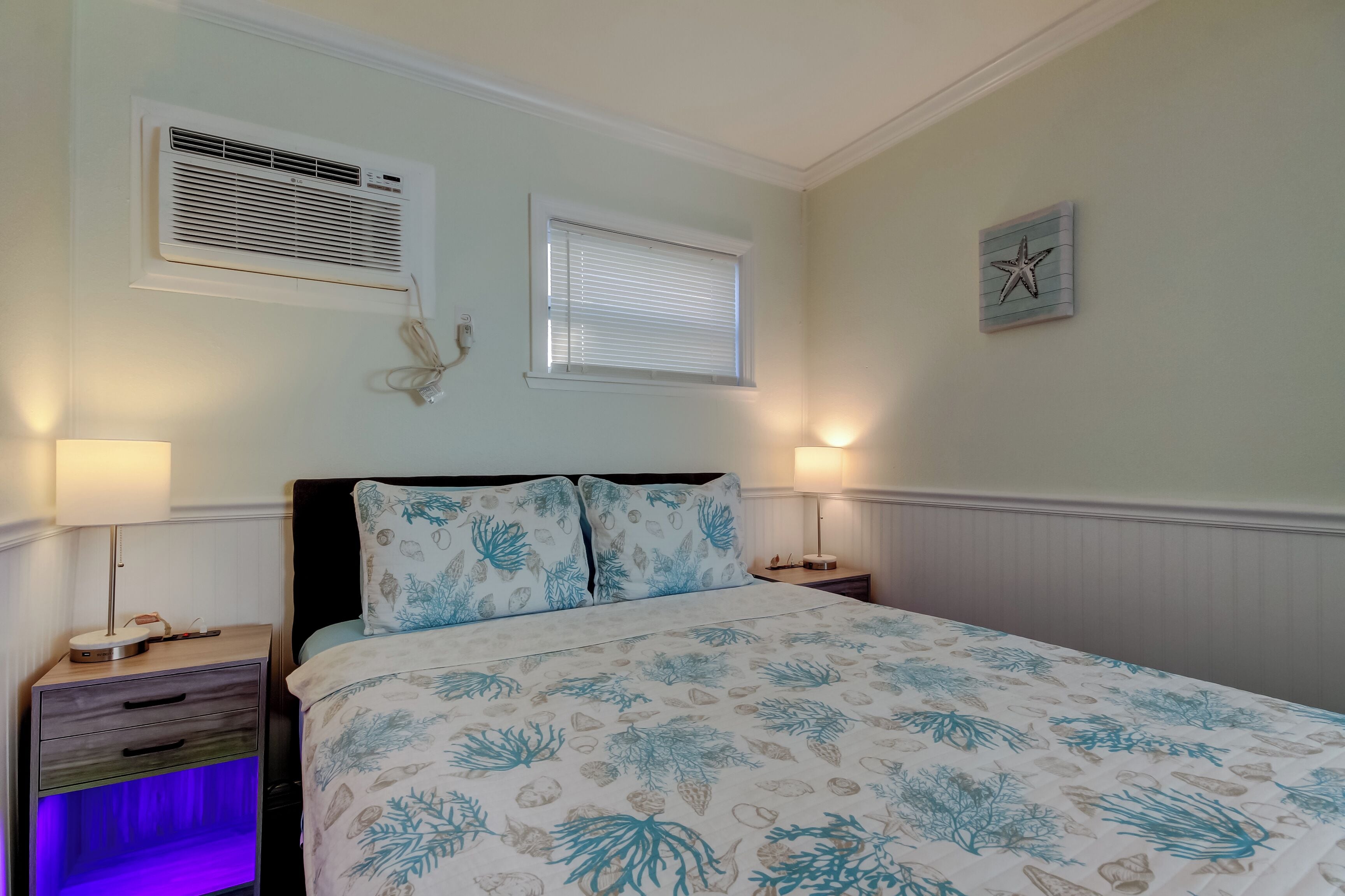 A bedroom with a bed, two bedside tables, a nightstand, a wall-mounted air conditioner, a window with blinds, and a wall decoration.