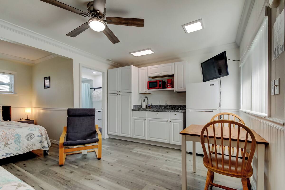 A compact room with a bed, a chair, a dining table, a ceiling fan, a TV, and a kitchenette.