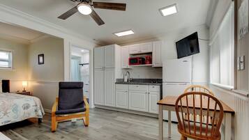 Una habitación pequeña con cama, silla, mesa de comedor, ventilador de techo, televisión y kitchenette.
