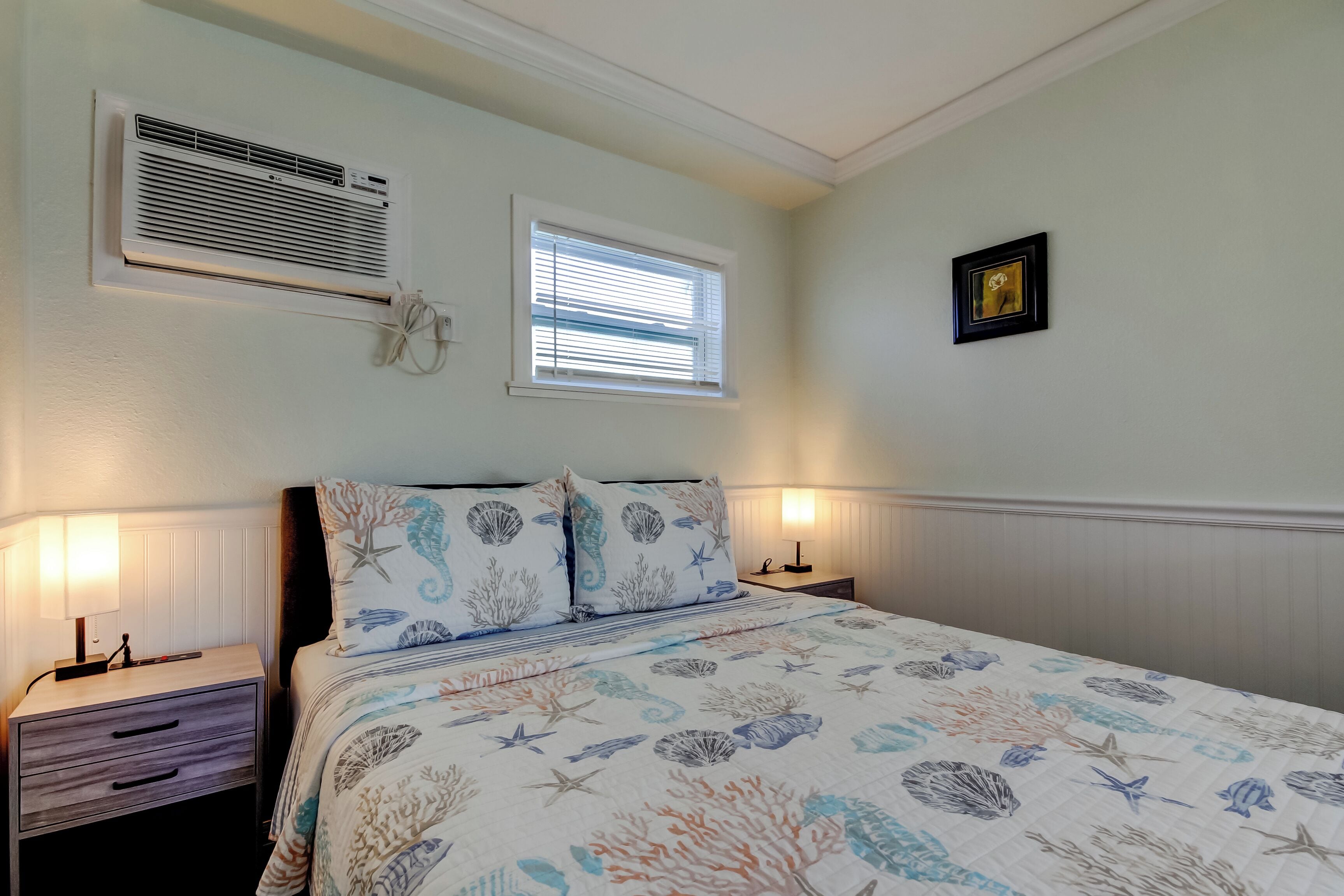 Une chambre à coucher comprenant un lit, deux tables de chevet, un climatiseur fixé au mur, une fenêtre avec des stores et un tableau encadré accroché au mur.