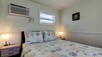 A bedroom with a bed, two bedside tables, a wall-mounted air conditioner, a window with blinds, and a framed picture on the wall.