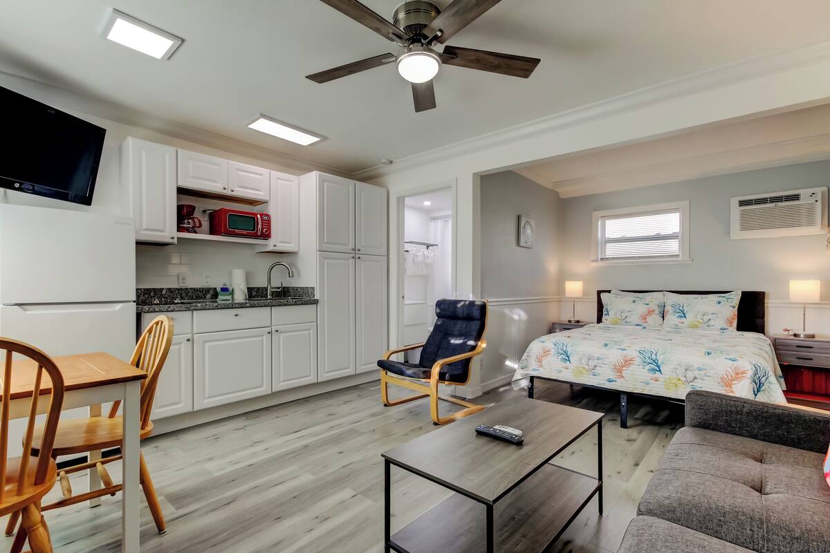 A compact living space with a kitchenette, a bed with a floral bedspread, a ceiling fan, and a television.