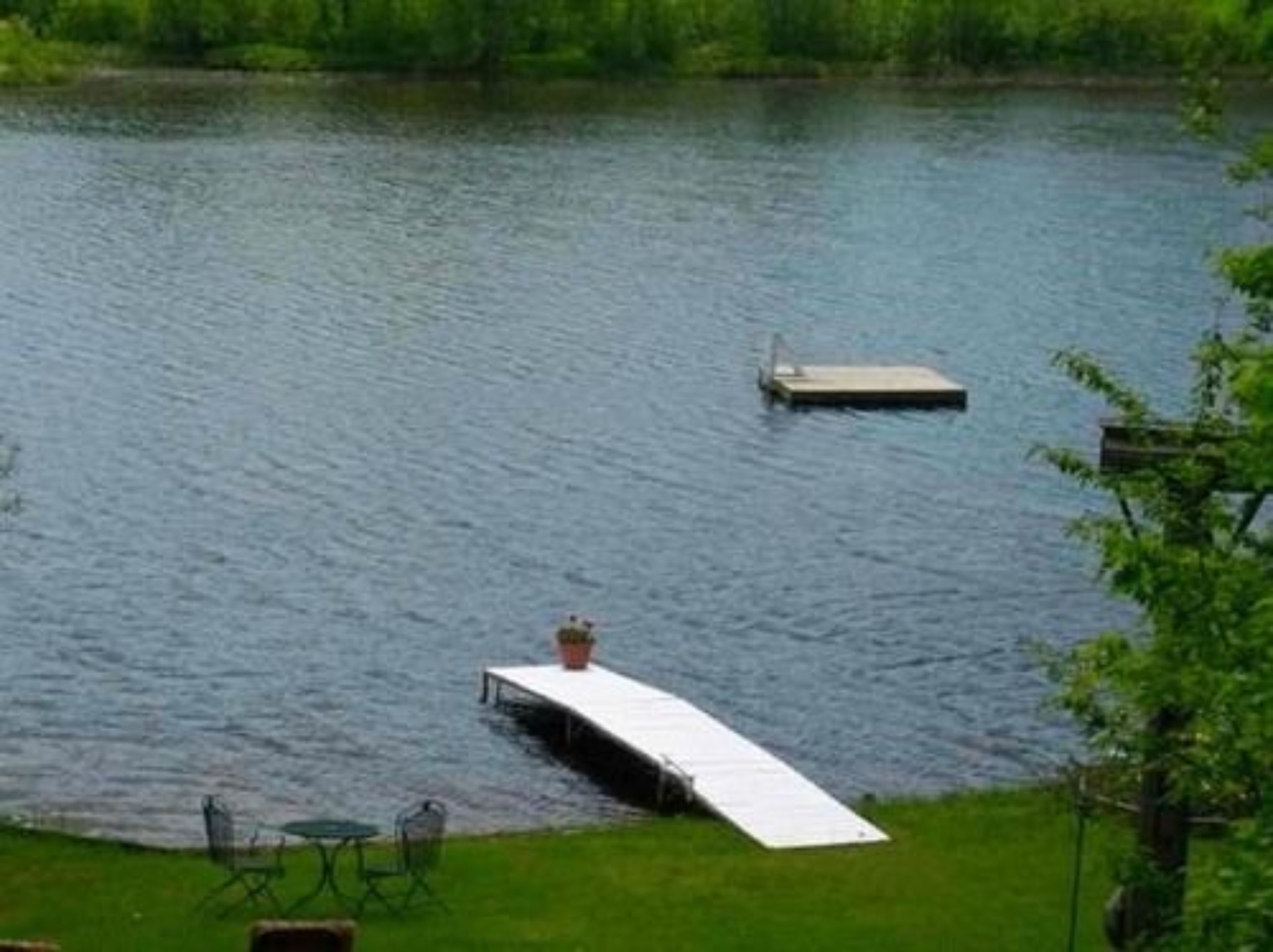 A lakeside view with a dock extending into the water, a grassy area with a table and chairs, and a small boat.