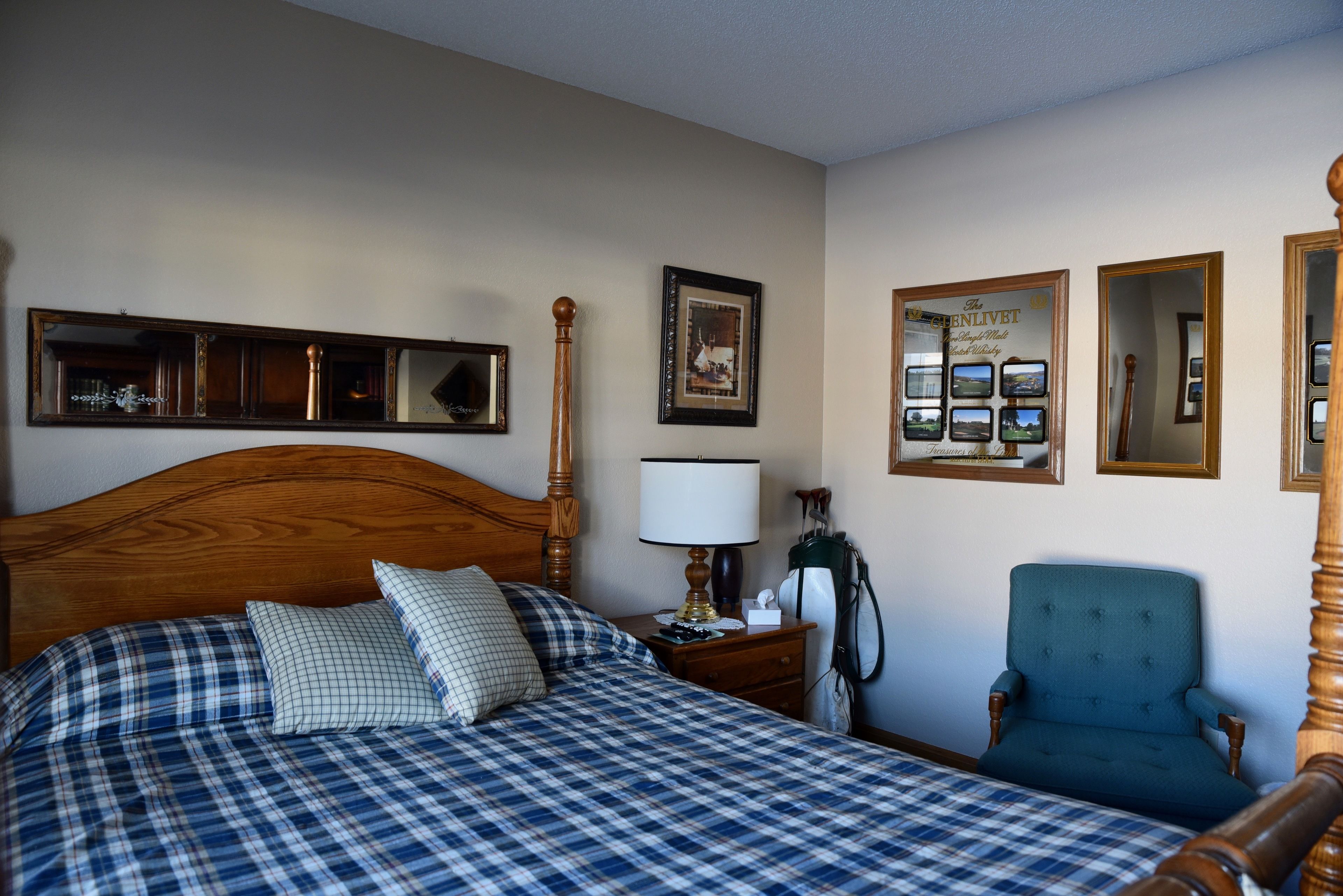 A bedroom with a wooden headboard, a bed with plaid bedding, a nightstand with a lamp, a chair, and framed pictures on the wall.