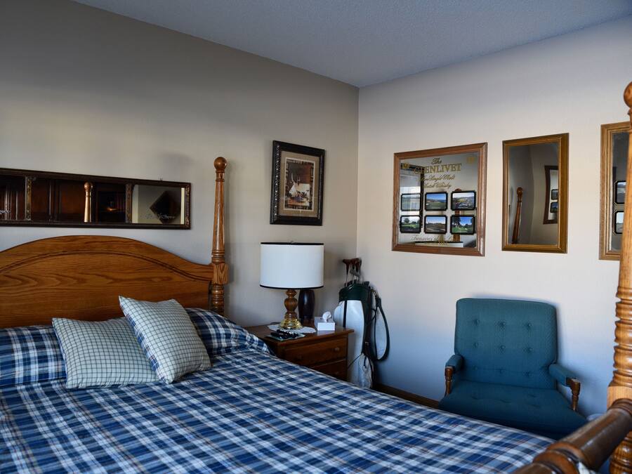 Une chambre à coucher avec une tête de lit en bois, un lit recouvert d’une couette à carreaux, une table de chevet avec une lampe, une chaise et des tableaux encadrés accrochés au mur.