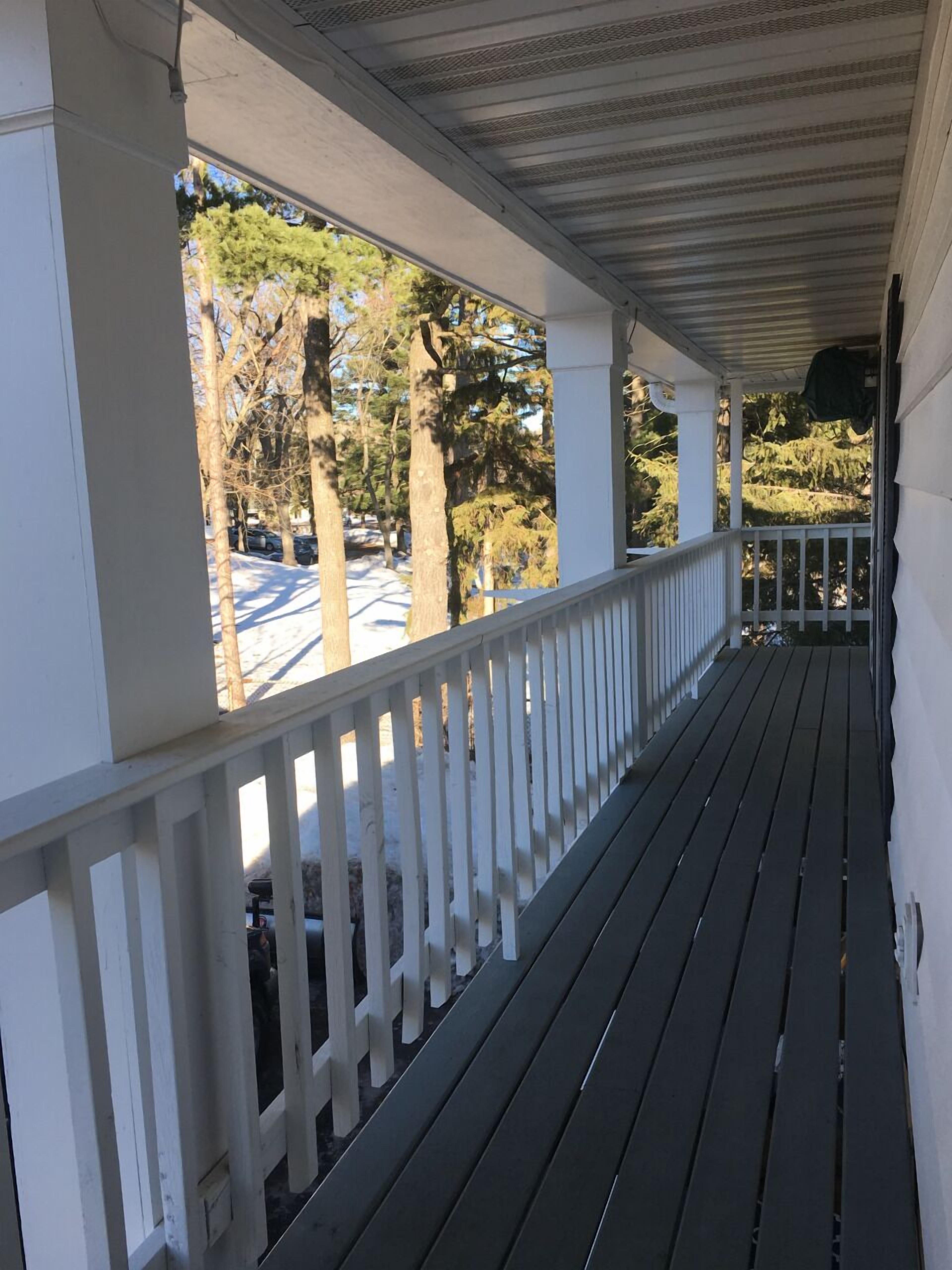 A covered porch with a white railing, wooden floor, and a view of trees and parked cars.