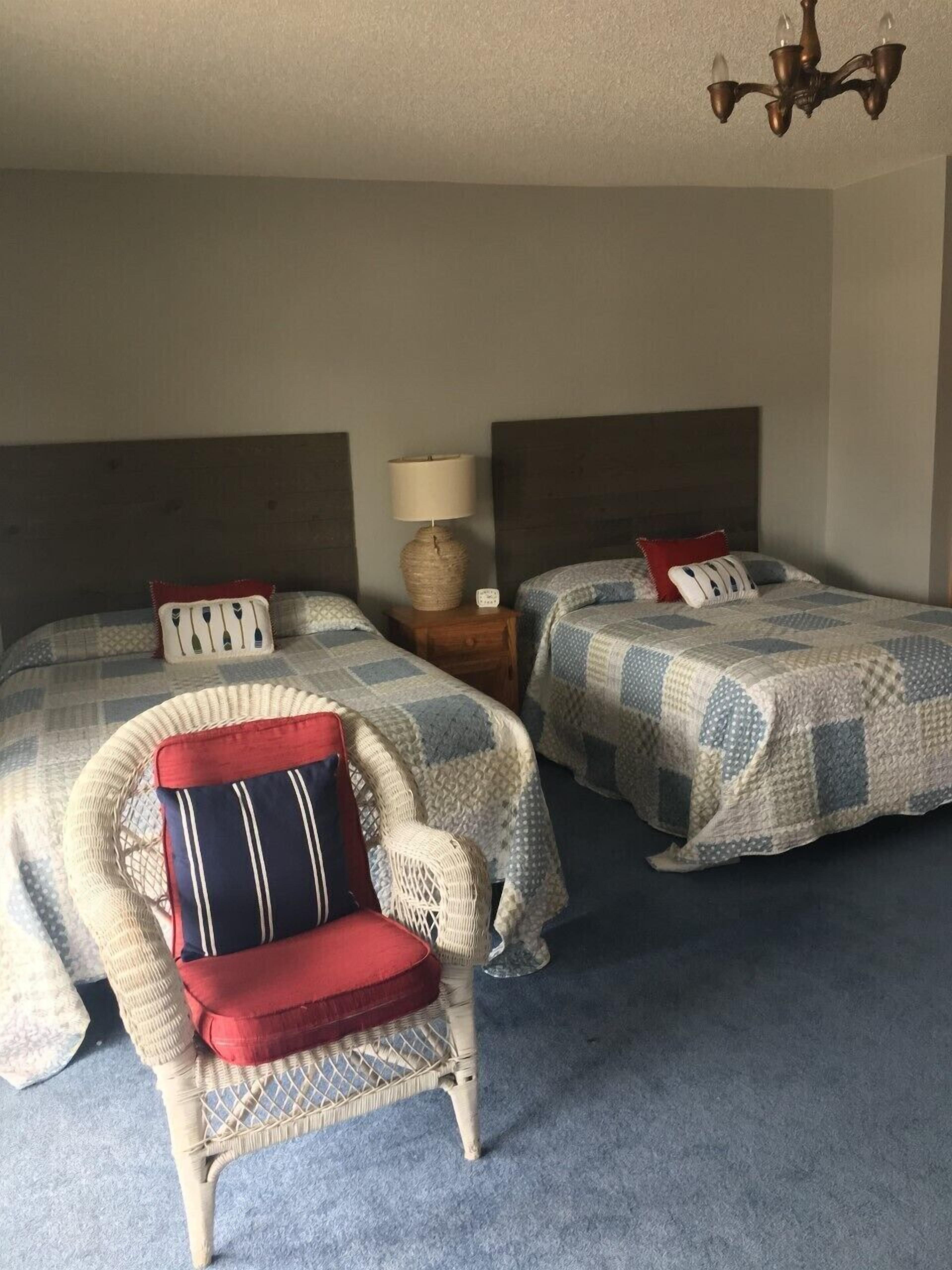 A hotel room with two beds, a wicker chair, a nightstand with a lamp, and a chandelier.